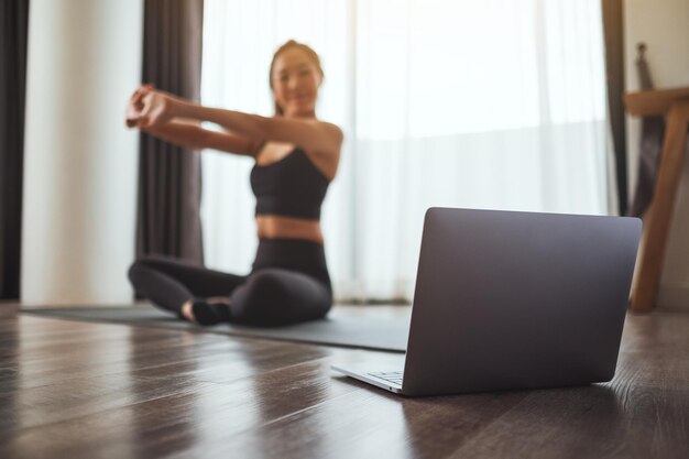 Une belle jeune femme asiatique étirant les bras tout en regardant des didacticiels d'entraînement en ligne sur un ordinateur portable à la maison