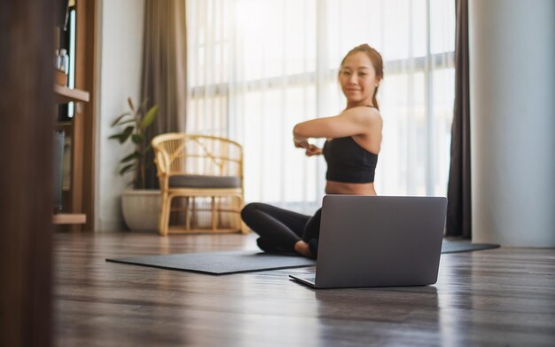 Une belle jeune femme asiatique étirant les bras tout en regardant des didacticiels d'entraînement en ligne sur un ordinateur portable à la maison