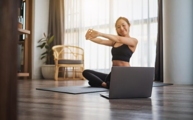 Une belle jeune femme asiatique étirant les bras tout en regardant des didacticiels d'entraînement en ligne sur un ordinateur portable à la maison