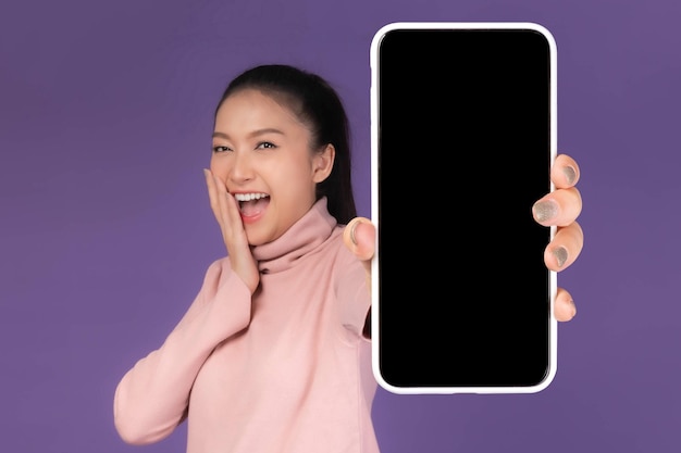 Belle jeune femme asiatique Fille surprise excitée montrant un grand téléphone intelligent avec écran blanc écran blanc isolé sur fond violet Afficher l'image simulée