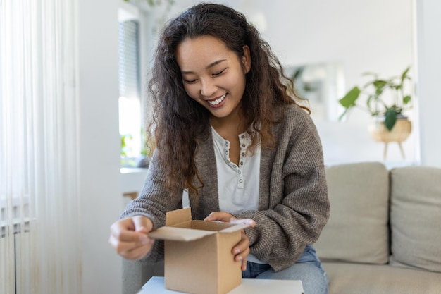 Belle jeune femme asiatique joyeuse déballant un paquet dans le salon d'un nouveau cadeau