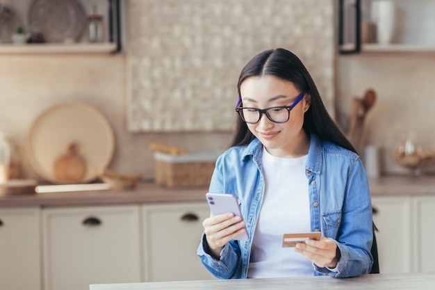 Une belle jeune femme asiatique à lunettes et une chemise en jean tient un téléphone et une carte de crédit en elle