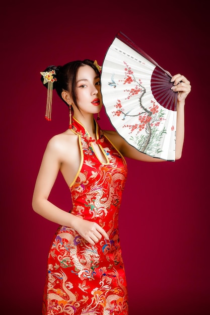 Belle jeune femme asiatique avec une peau fraîche et propre vêtue d'une robe traditionnelle cheongsam qipao tenant un ventilateur posant sur fond rouge Portrait de modèle féminin en studio Joyeux nouvel an chinois