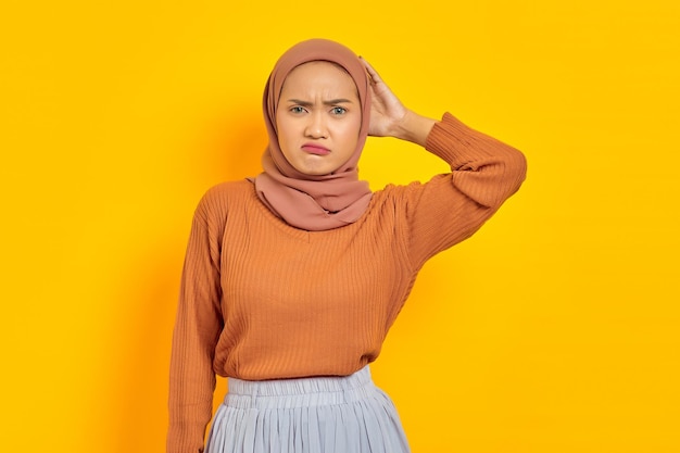 Belle jeune femme asiatique en pull marron et hijab confus à propos d'une question, pensant avec la main sur la tête isolée sur fond jaune. Concept religieux de l'islam des gens
