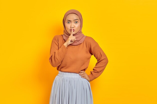 Belle jeune femme asiatique en pull marron et hijab montrant un geste de silence avec le doigt et regardant la caméra isolée sur fond jaune. Concept religieux de l'islam des gens