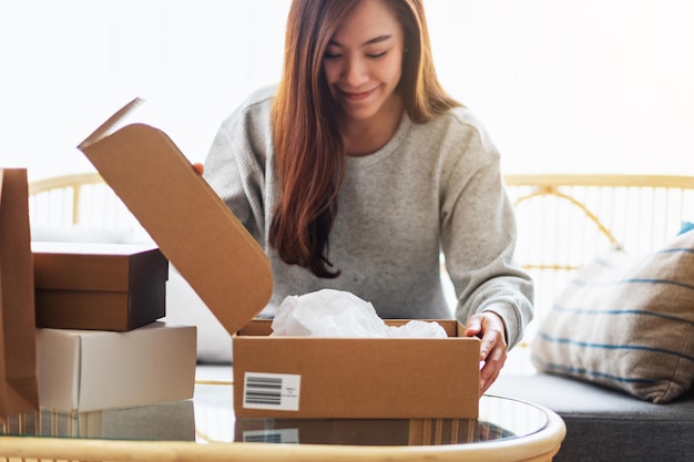 Une belle jeune femme asiatique recevant et ouvrant une boîte à colis postal à la maison pour la livraison et le concept d'achat en ligne