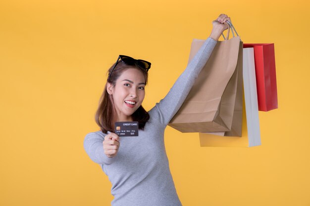 Belle jeune femme asiatique avec des sacs à provisions colorés et carte de crédit isolé sur mur jaune