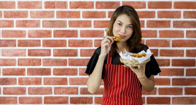 Belle jeune femme asiatique séduisante en tablier rayé rouge debout près d'un mur de briques et persuade en souriant d'apprécier de manger de la nourriture délicieuse préférée en levant une collation frite et son plateau près de la bouche