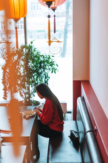 Belle jeune femme asiatique souriante en rouge mangeant de la nourriture asiatique avec des baguettes en bambou au restaurant