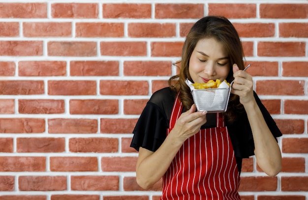 Belle jeune femme asiatique en tablier rayé rouge tenant un tamis de délicieuses pommes de terre frites près d'un mur de briques et le montre fièrement avec le sourire comme satisfaisant ses compétences culinaires et persuade de manger de la nourriture délicieuse