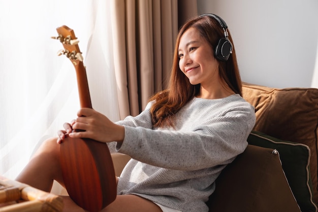 Une belle jeune femme asiatique tenant et regardant un ukulélé tout en écoutant de la musique avec un casque à la maison