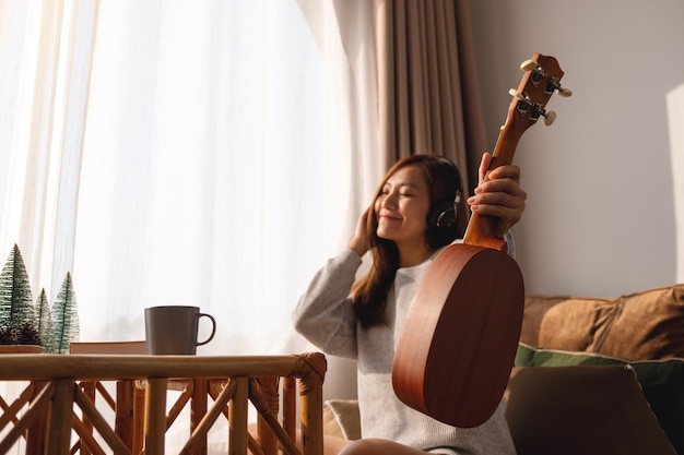 Une belle jeune femme asiatique tenant un ukulélé et aime écouter de la musique avec un casque à la maison