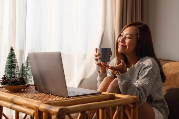 Une belle jeune femme asiatique utilisant un ordinateur portable pour un appel vidéo assis et buvant du café à la maison