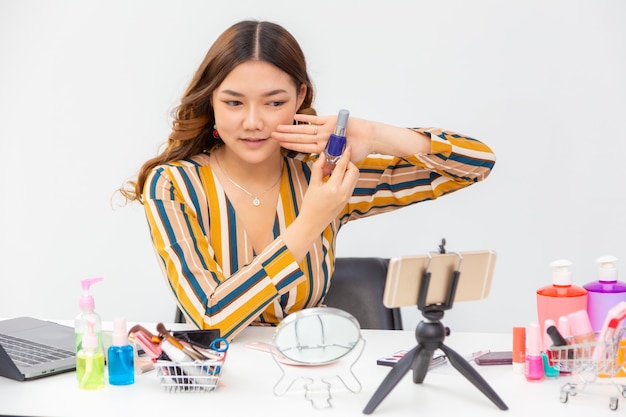 Belle jeune femme asiatique, vlogger, montrant et examinant des produits de beauté sur un blog vidéo à la maison en utilisant son téléphone pour diffuser en direct