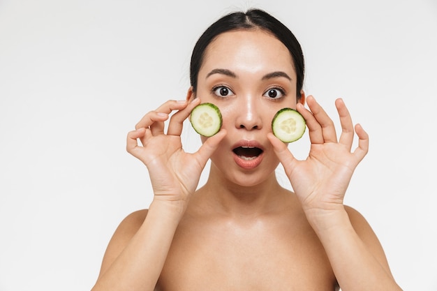 belle jeune femme assez asiatique avec une peau saine posant nue isolée sur un mur blanc tenant du concombre.