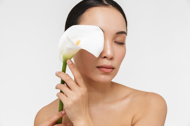 belle jeune femme assez asiatique avec une peau saine posant nue isolée sur un mur blanc tenant une fleur.