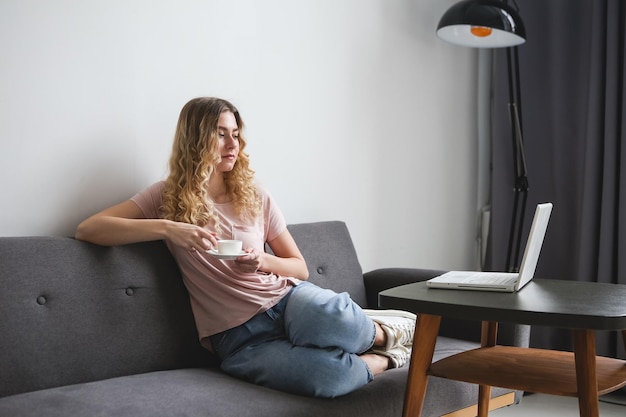 Photo belle jeune femme assise sur le canapé avec une tasse de café blanche travaillant sur un ordinateur portable blanc