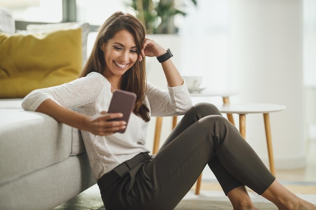 Belle jeune femme assise à côté de la fenêtre, ayant du temps libre, tenant un téléphone intelligent et surfant sur les réseaux sociaux chez elle.