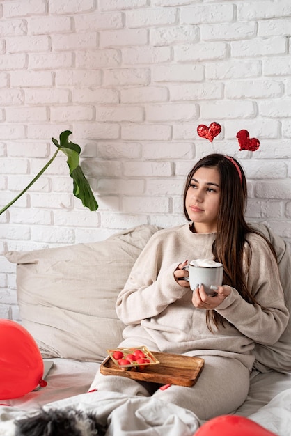 Belle jeune femme assise dans le lit célébrant la saint valentin mangeant des bonbons et buvant du café