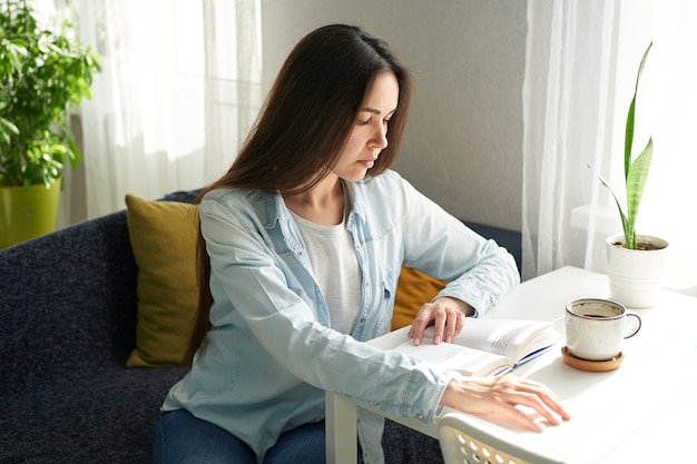 Belle jeune femme assise à la table à la maison et lisant un livre