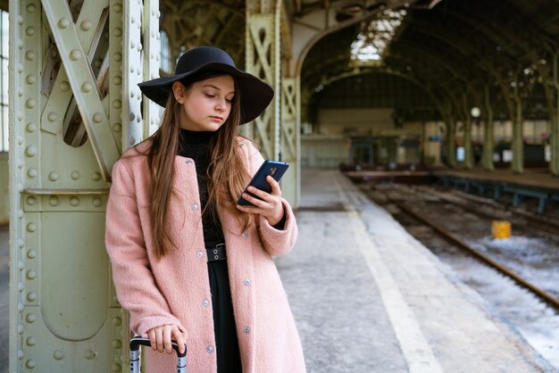 Belle jeune femme attendant le train à la gare pour voyager en automne avec le téléphone à la main il ...