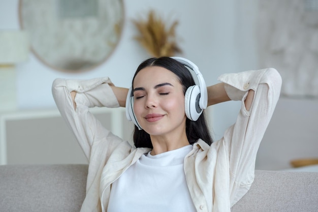 Belle jeune femme au casque assis sur le canapé à la maison avec les yeux fermés et les mains derrière