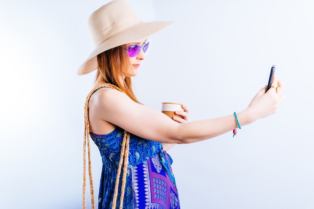 Belle jeune femme au chapeau d'été tasse de café et robe prenant un selfie sur fond blanc