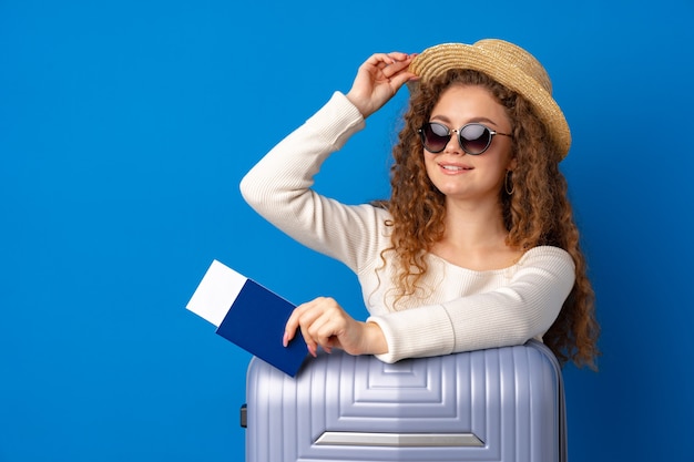 Belle jeune femme au chapeau en vacances avec valise sur fond bleu