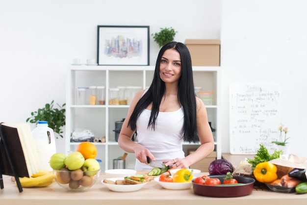 Belle jeune femme au foyer dans la cuisine, coupe de légumes verts frais, souriant, regardant la caméra. Livre de cuisine ouvert avec des recettes sur la table de travail du cuisinier.