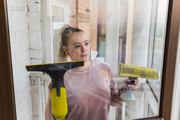 Belle jeune femme au foyer en vêtements de travail lavant les vitres par des outils professionnels à la maison.