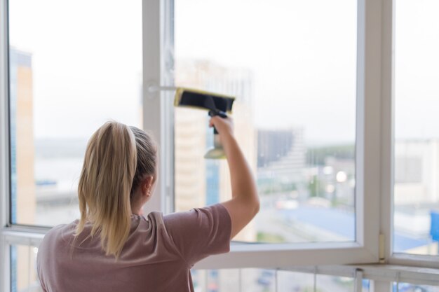 Belle jeune femme au foyer en vêtements de travail lavant les vitres par des outils professionnels à la maison.