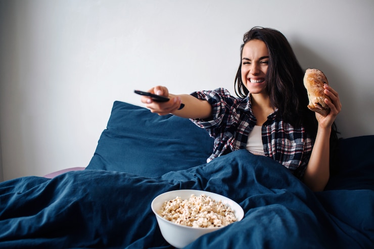 Photo belle jeune femme au lit du matin à la maison. femme heureuse joyeuse devant la télé. tenez la télécommande en main et utilisez-la. petit déjeuner avec pop-corn et sandwich.