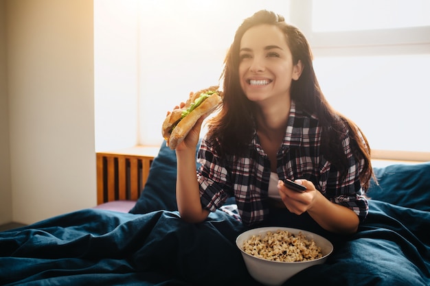 Belle jeune femme au lit du matin à la maison. Regardez la télévision ou un film et utilisez la télécommande. Sandwich et pop-corn.