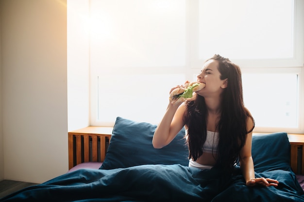 Belle jeune femme au lit du matin à la maison. Sandwich manger et mordre avec des légumes verts et du pain savoureux. Petit déjeuner à la maison.