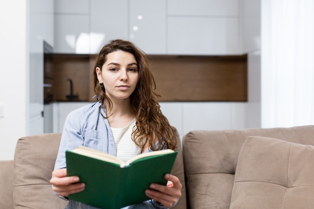 Belle jeune femme au repos à la maison assis sur le canapé et lisant un livre intéressant