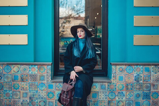Belle jeune femme aux cheveux bleus dans un joli manteau noir, un jean et un chapeau.
