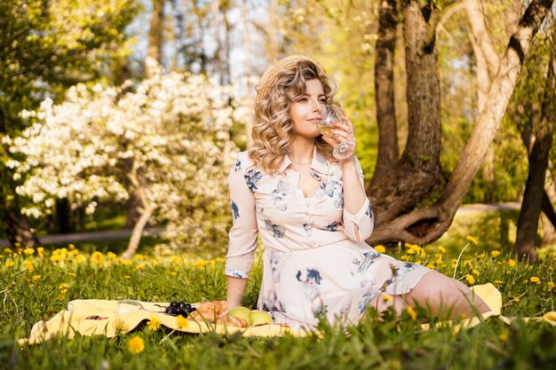 Belle jeune femme aux cheveux blonds en chapeau de paille boit du vin et assis sur le plaid dans le jardin lors d'un pique-nique d'été