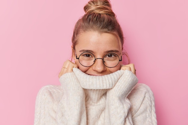 Belle jeune femme aux cheveux blonds rassemblés en chignon cache la bouche derrière le col du pull porte des lunettes rondes isolées sur fond rose. Un modèle féminin ravi et sincère porte un pull chaud