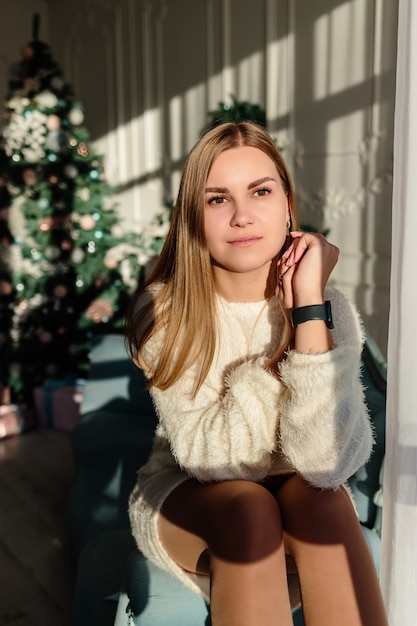 Une belle jeune femme aux cheveux blonds se dresse sur fond d'arbre de Noël. Le nouvel an arrive bientôt. Ambiance de Noël dans une maison cosy