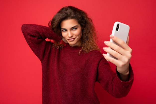 Belle jeune femme aux cheveux bouclés portant un pull rouge foncé isolé sur le mur de fond rouge