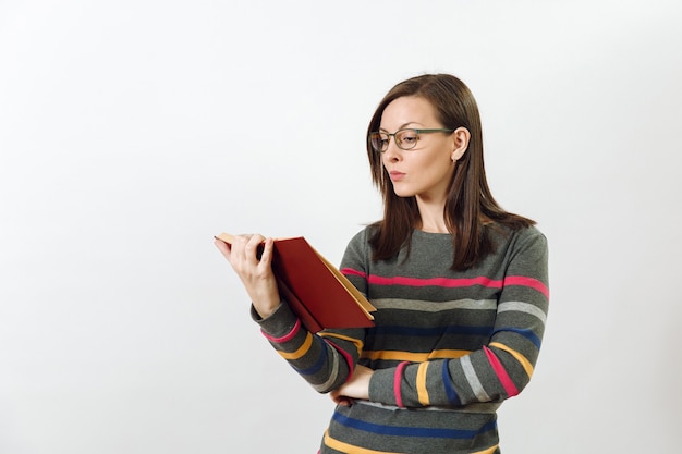 Une belle jeune femme aux cheveux bruns européenne dans des verres pour la vue vêtue d'une chemise à manches longues gris foncé décontractée avec des rayures colorées, debout avec un livre sur fond blanc. Concept de lecture et d'étude