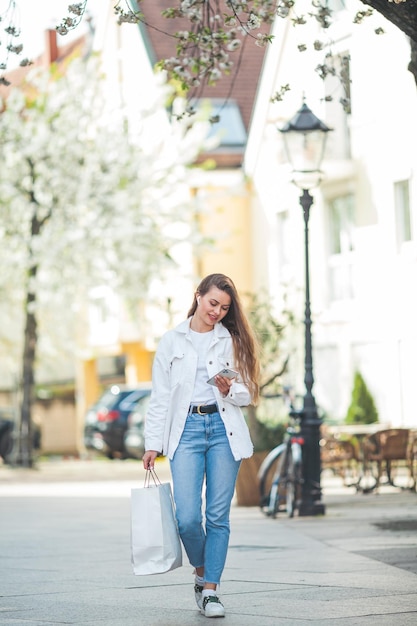 Une belle jeune femme aux cheveux longs dans des vêtements décontractés légers et confortables écoute de la musique à l'aide d'un casque Bluetooth se promène dans le centre d'une ville européenne Un joli modèle fait du shopping au printemps