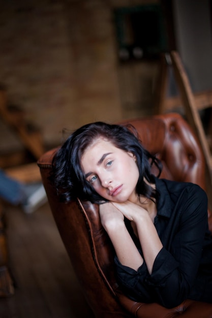 Belle jeune femme aux cheveux noirs bouclés et au maquillage naturel assise sur des chaises en cuir foncé