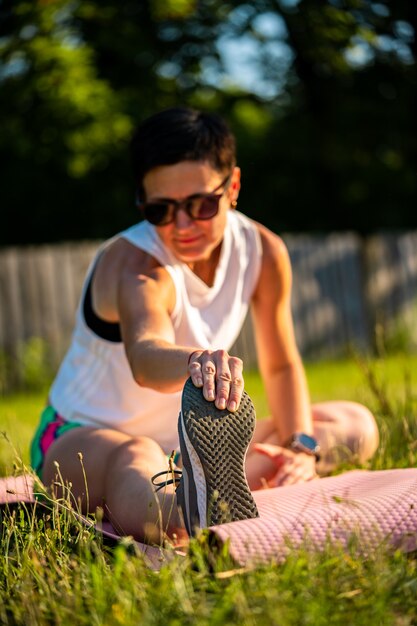 Belle jeune femme aux cheveux noirs courts s'étendant sur un tapis de yoga sur une herbe dans un parc, se réchauffe avant l'entraînement