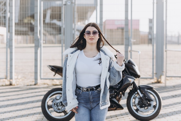 Belle jeune femme aux cheveux noirs en pantalon en jean et une veste près d'une moto de sport noire