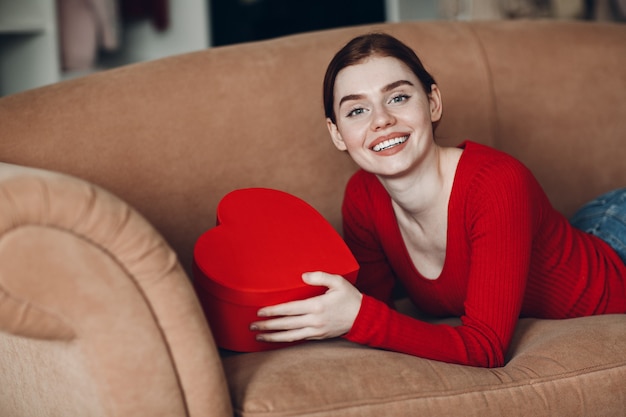 Belle jeune femme aux cheveux rouges allongée dans son canapé dans le salon et tenant le coffret cadeau en forme de coeur et de sourire. Saint Valentin ou anniversaire