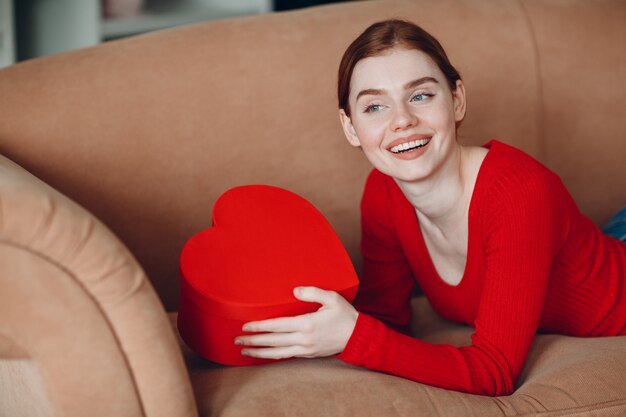 Belle jeune femme aux cheveux rouges allongée dans son canapé dans le salon et tenant le coffret cadeau en forme de coeur et de sourire. Saint Valentin ou anniversaire