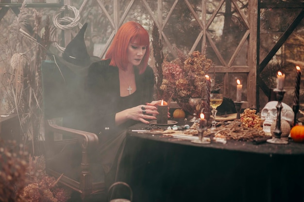 Photo belle jeune femme aux cheveux rouges en chapeau noir et tenue de sorcière fait de la magie dans une serre décorée pour la fête d'halloween avec des citrouilles et un crâne dans la fumée