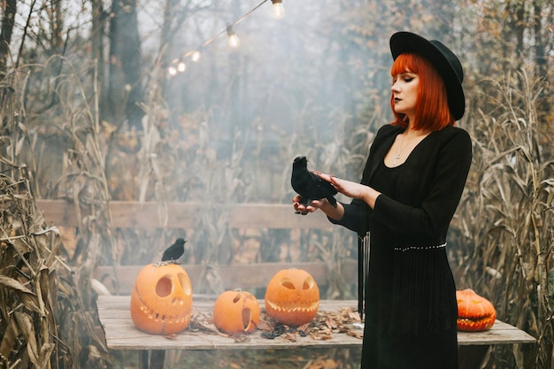 Belle jeune femme aux cheveux rouges en chapeau noir et tenue de sorcière s'amuse dans la cour arrière décorée pour la fête d'Halloween avec des citrouilles et un corbeau noir dans la fumée