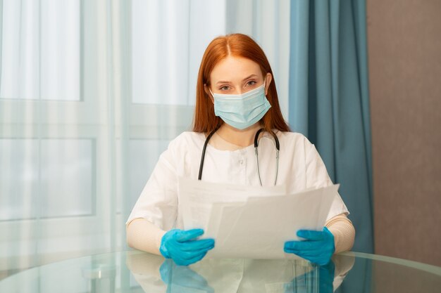 Belle jeune femme aux cheveux rouges dans une robe médicale et un masque